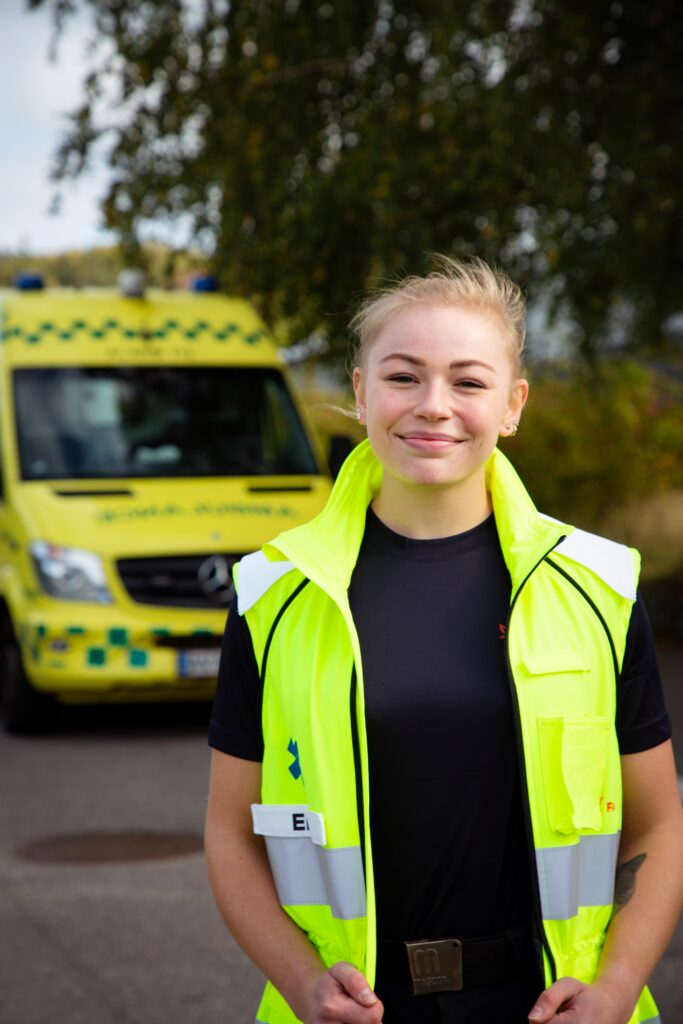 Smilende ung ambulancebehandler i refleksvest står foran Mercedes-ambulance.