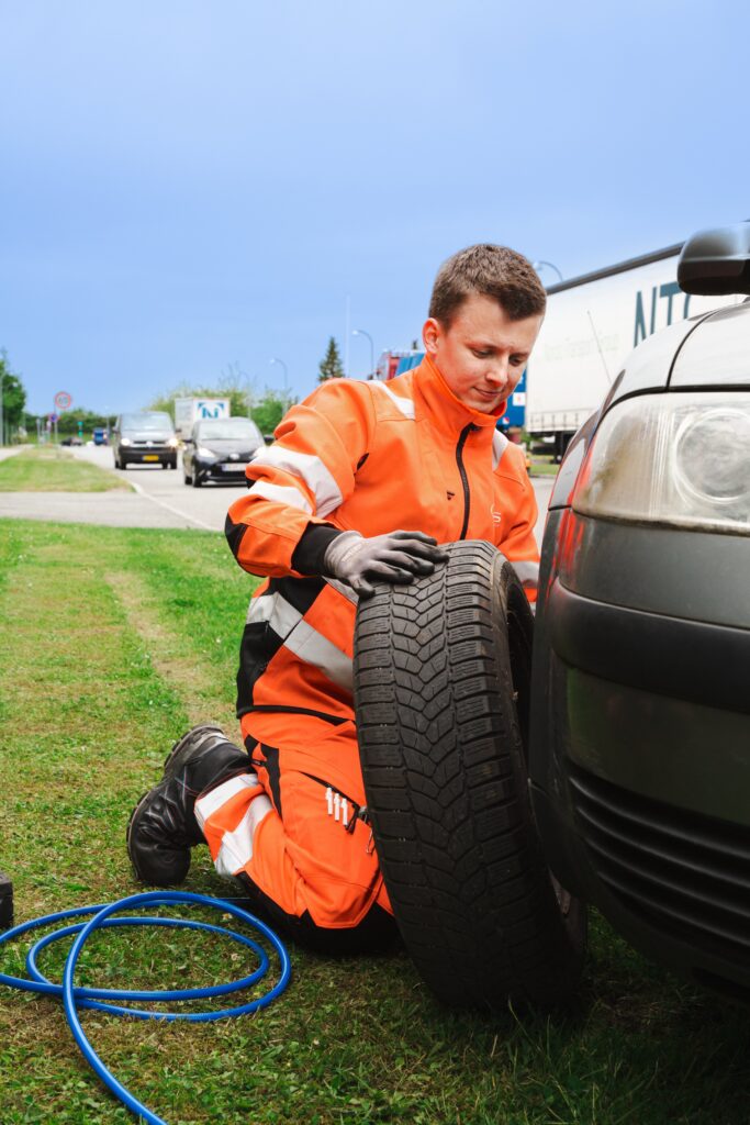 Autohjælper knæler ved vejkanten og skifter dæk på en bilist langs landevejen.