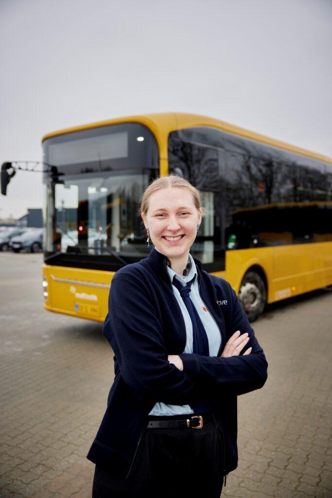 Smilende buschauffør i Umove-uniform står foran sin gule Midttrafik-bybus.