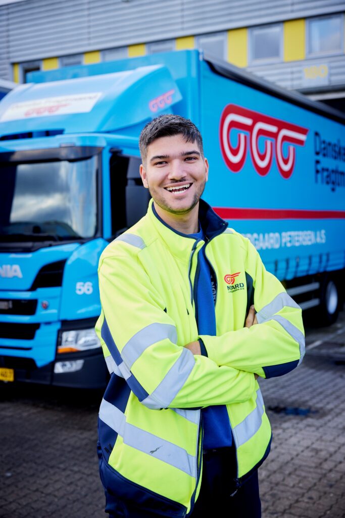 Portræt af glad godschauffør i refleksjakke foran Danske Fragtmænd Scania-lastbil.