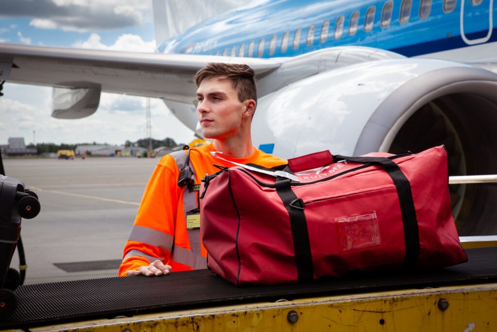 Lufthavnsoperatør læsser bagage på transportbånd foran et KLM-fly.